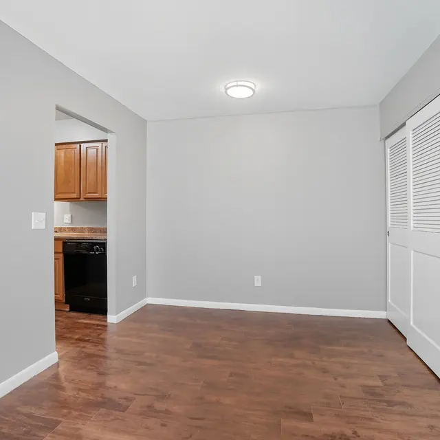 A spacious, empty room with gray walls and brown wooden flooring. There is a closet with white bi-fold doors on the right and an opening to a kitchen area visible on the left.