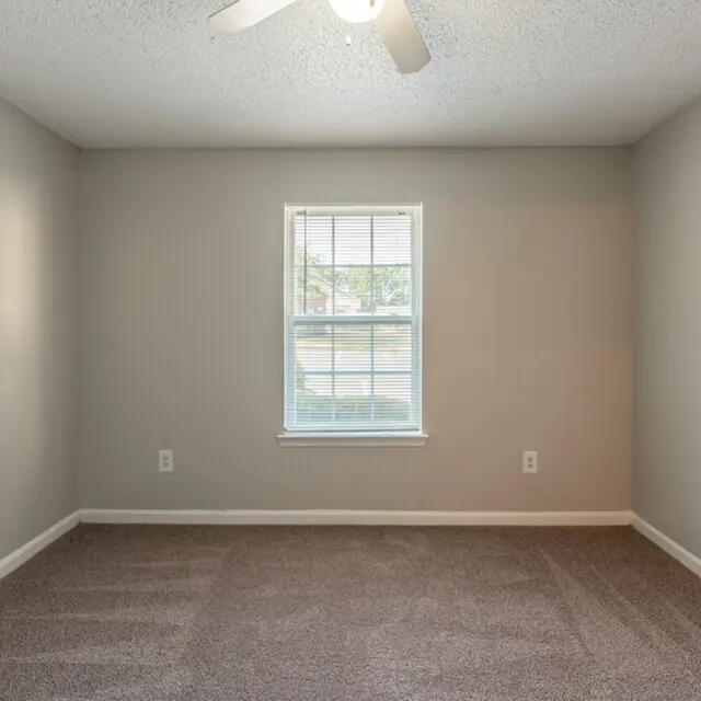 An empty, well-lit room featuring a textured ceiling and a ceiling fan. The walls are painted a light gray, and the floor is covered in soft brown carpet. A window with white blinds allows natural light to enter the room.