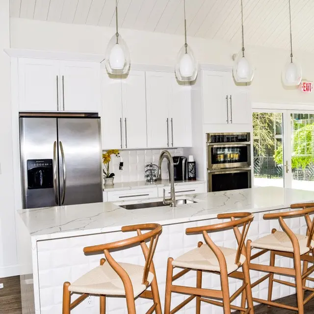 A modern kitchen with a large island, white cabinetry, stainless steel appliances, and wooden bar stools. Natural light floods the space through large windows