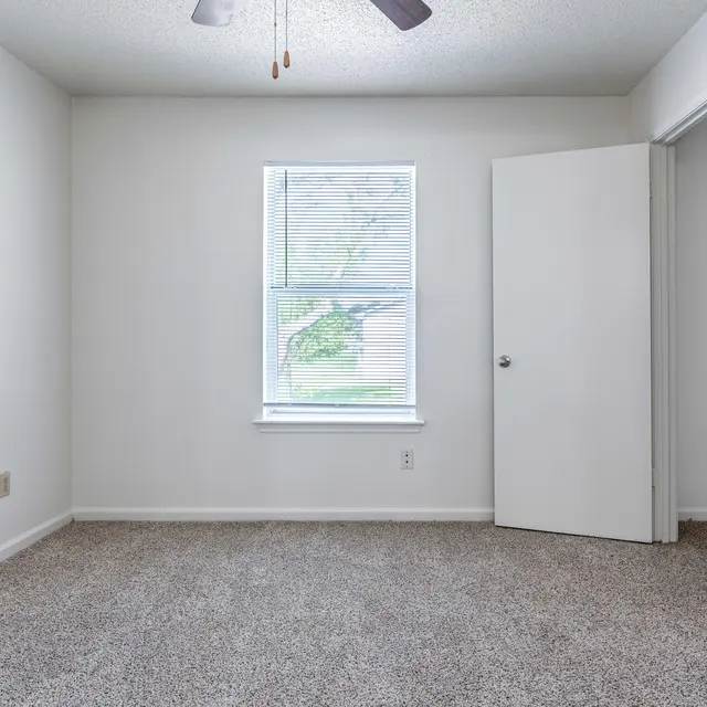 A spacious, empty room with beige carpet, a ceiling fan, and a window with blinds. A partially open white door is visible on the right side, leading to another room.