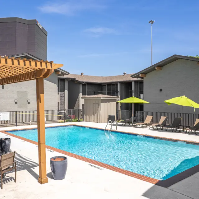 A bright outdoor pool area at a residential complex featuring a blue swimming pool surrounded by lounge chairs and green umbrellas.