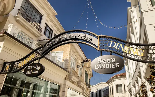 A decorative archway sign featuring the words 'Dining' and 'See's Candies,' with additional signage for 'Brandy Melville.' The backdrop shows a bright blue sky and buildings.