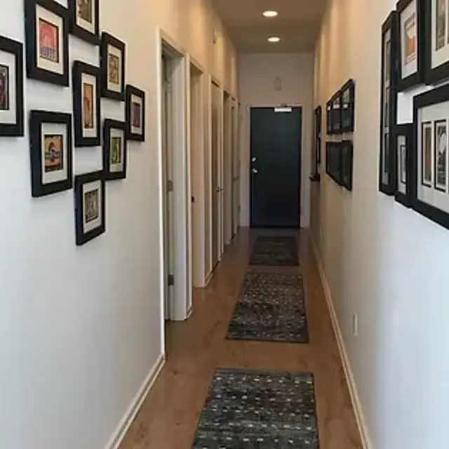 A long, narrow hallway with white walls, featuring framed pictures on the left side and a blue door at the end. There are patterned rugs lining the wooden floor.