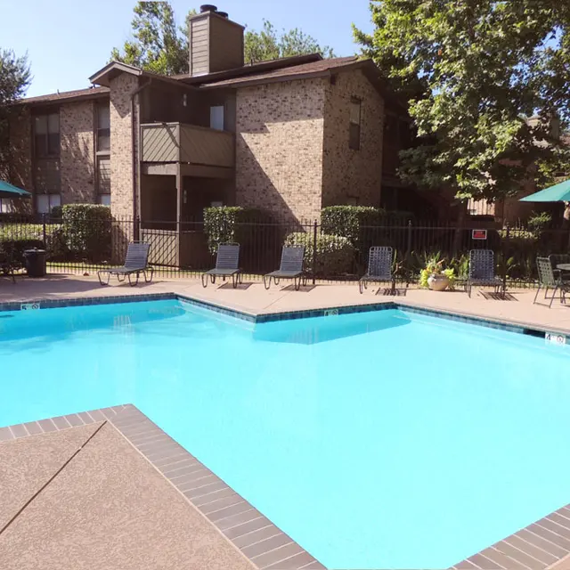 A sunny outdoor swimming pool surrounded by lounge chairs and umbrellas at an apartment complex.