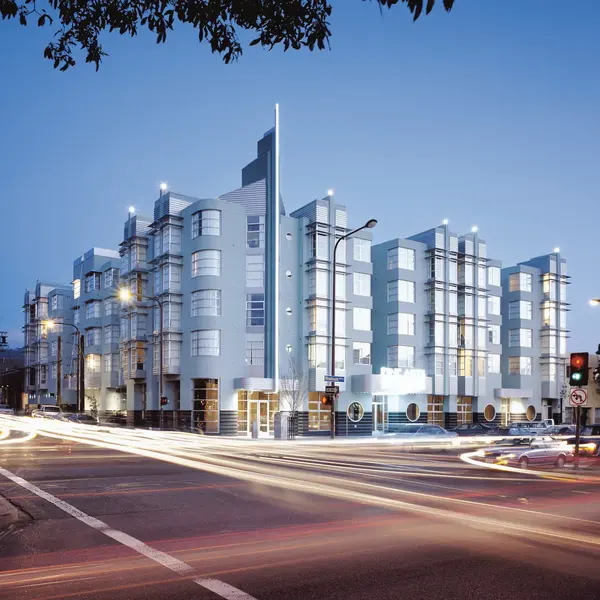 A modern multi-story residential building viewed from an intersection, featuring a blend of architectural styles with a sleek facade and large windows, illuminated by streetlights at dusk.