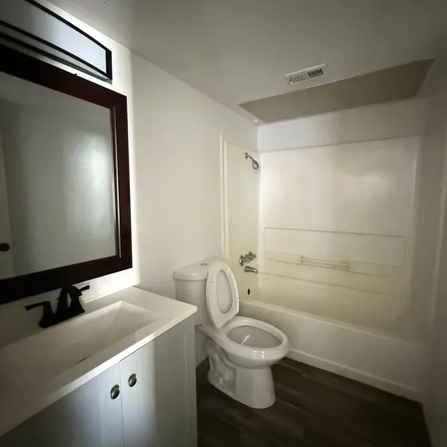A clean and modern bathroom featuring a white vanity with a mirror, a toilet, and a bathtub with a shower. The flooring is a dark wood finish, creating a contemporary look.