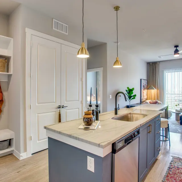 A stylish kitchen featuring a marble countertop island, with pendant lighting, an open layout leading to a cozy living area visible through a large window. The decor is modern, with a mixture of neutral tones and a touch of greenery from a nearby plant.