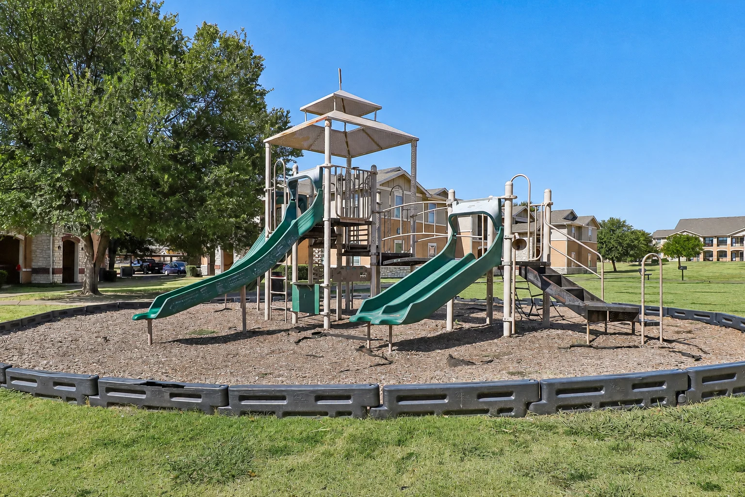 A playground with green slides and a structure under a clear blue sky, surrounded by grass.
