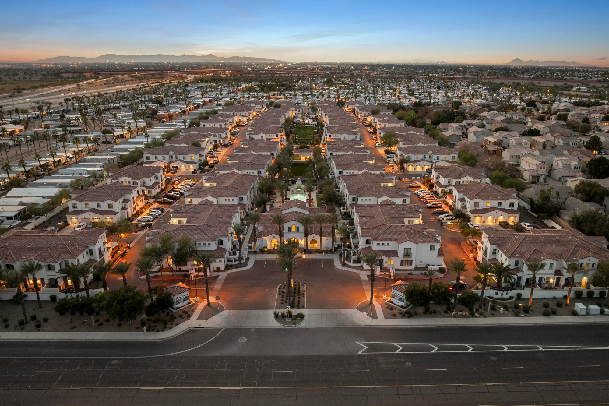 An aerial view of a residential neighborhood at dusk, showcasing rows of houses with terracotta roofs, palm trees, and well-lit streets.