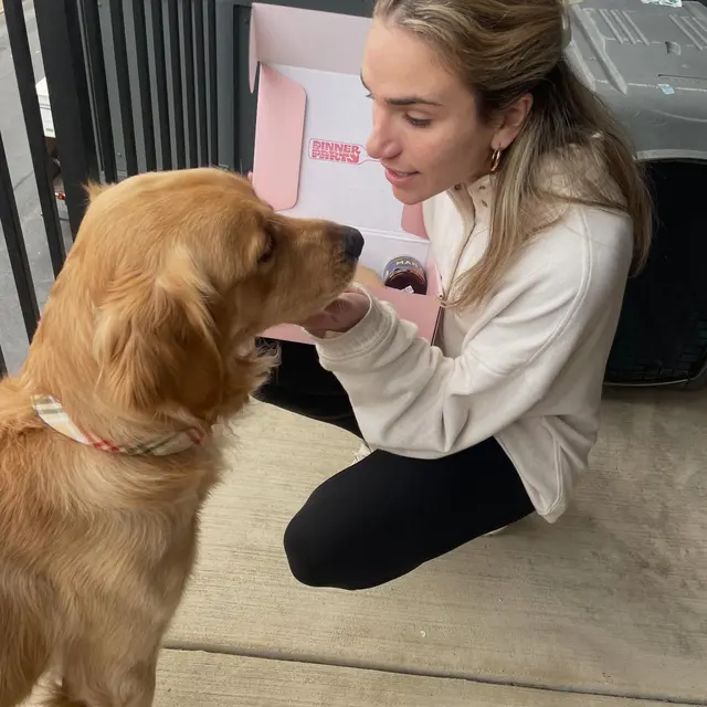 A woman kneeling on a porch, interacting with a golden retriever while a pink box is placed nearby.