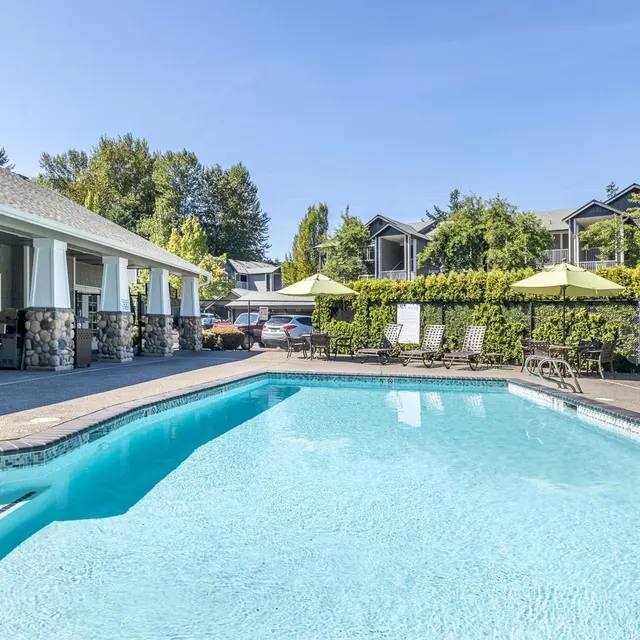 A clear blue swimming pool surrounded by lounge chairs and umbrellas, with a residential building in the background.