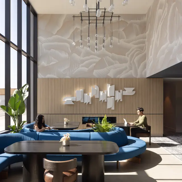 A modern lobby interior featuring large windows with natural light. The space includes blue curved seating, a dark wooden table, and decorative plants. Abstract wall art is displayed above the seating area, and a contemporary chandelier hangs from the ceiling. Two people are seated, one reading and the other working on a laptop.