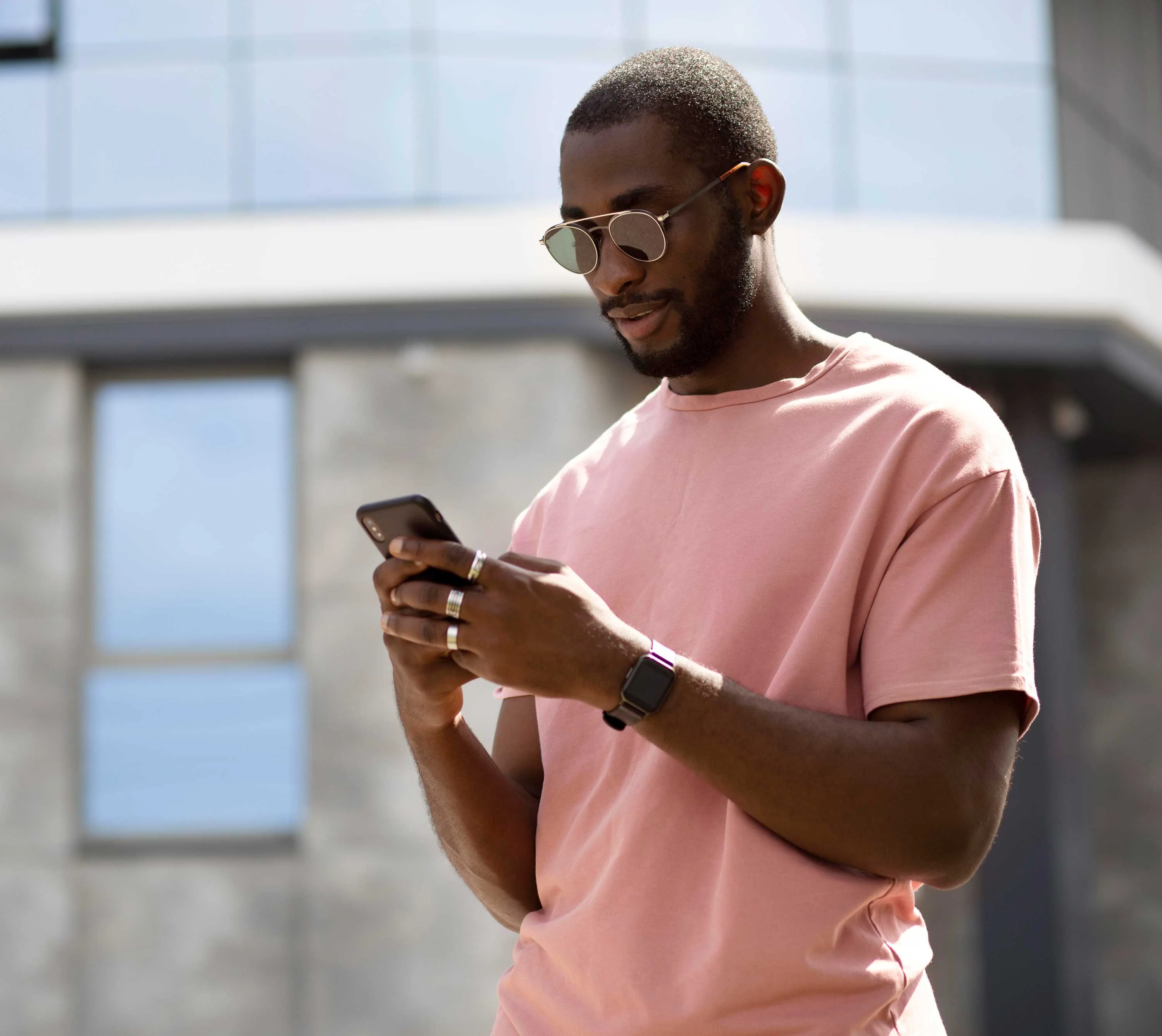 A man wearing sunglasses and a pink t-shirt checks his phone while standing outdoors. The background features a modern building with large windows.