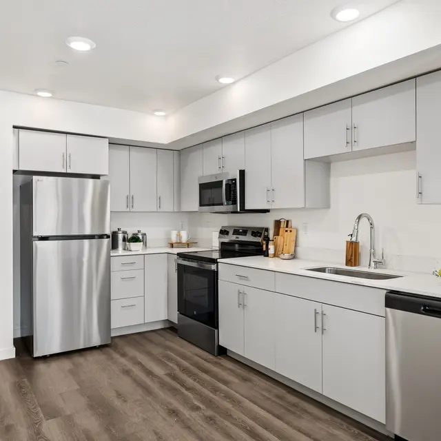 A sleek modern kitchen featuring stainless steel appliances, light gray cabinetry, and wooden flooring. The kitchen has a refrigerator, oven, and sink with an island countertop. A bowl of yellow fruit decorates the countertop.
