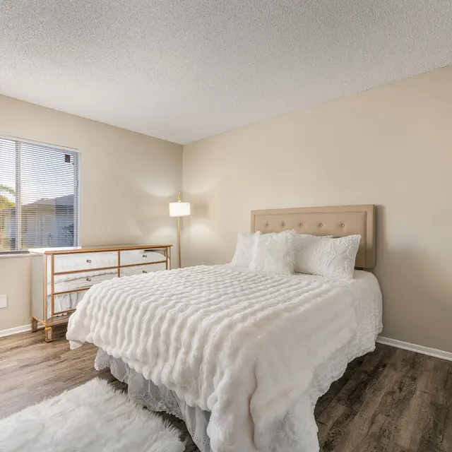 A modern bedroom featuring a queen-sized bed with a plush white comforter, a bedside lamp, and a window with blinds allowing natural light to enter. A wooden dresser is positioned next to the bed on a wooden floor.