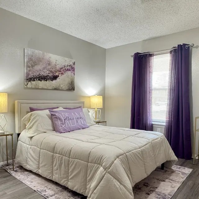 A cozy bedroom featuring a bed with a light-colored duvet, purple accent pillows, and framed artwork on the wall. The room has two bedside lamps, a window with purple curtains, and a decorative mirror. A small side table and chair are also present.