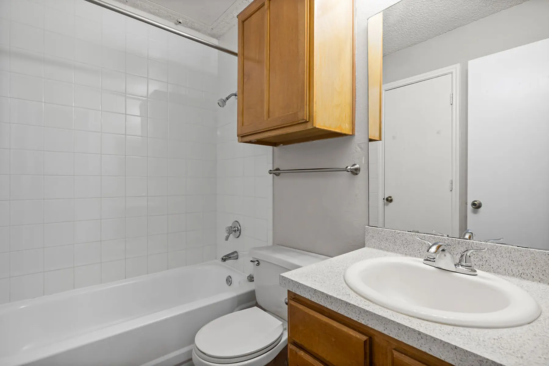 Simple Bathroom Interior A clean and simple bathroom featuring a white bathtub, toilet, and sink with a granite countertop. There are wooden cabinets above the sink, and the walls are tiled in a white color. A mirror reflects the bathroom space, and there is a door leading out of the bathroom.