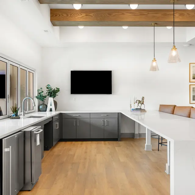 A modern kitchen with gray cabinetry, a large white countertop, and wooden beams on the ceiling. It features a flat-screen TV on the wall, pendant lighting, and a seating area with brown bar stools.