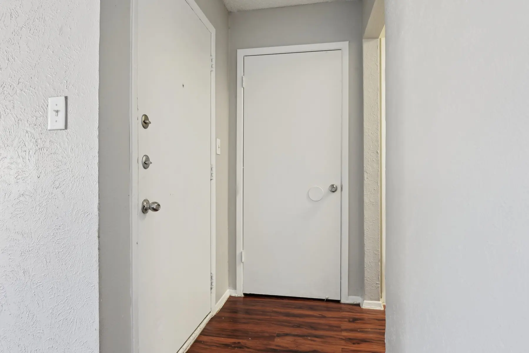A view of a hallway featuring a white front door and a closed white interior door, with wooden flooring and light gray walls.