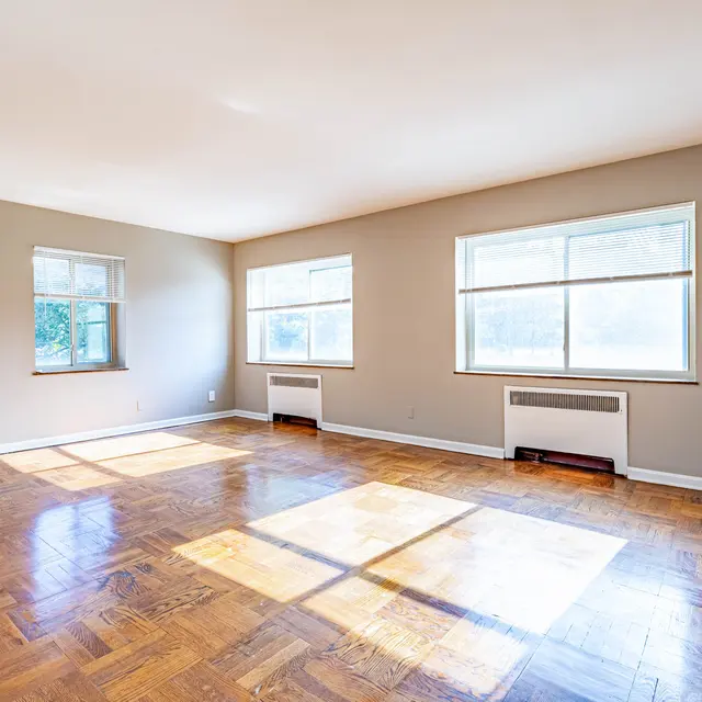 An empty living room with hardwood floors, featuring two large windows providing natural light, and a wall-mounted air conditioning unit.