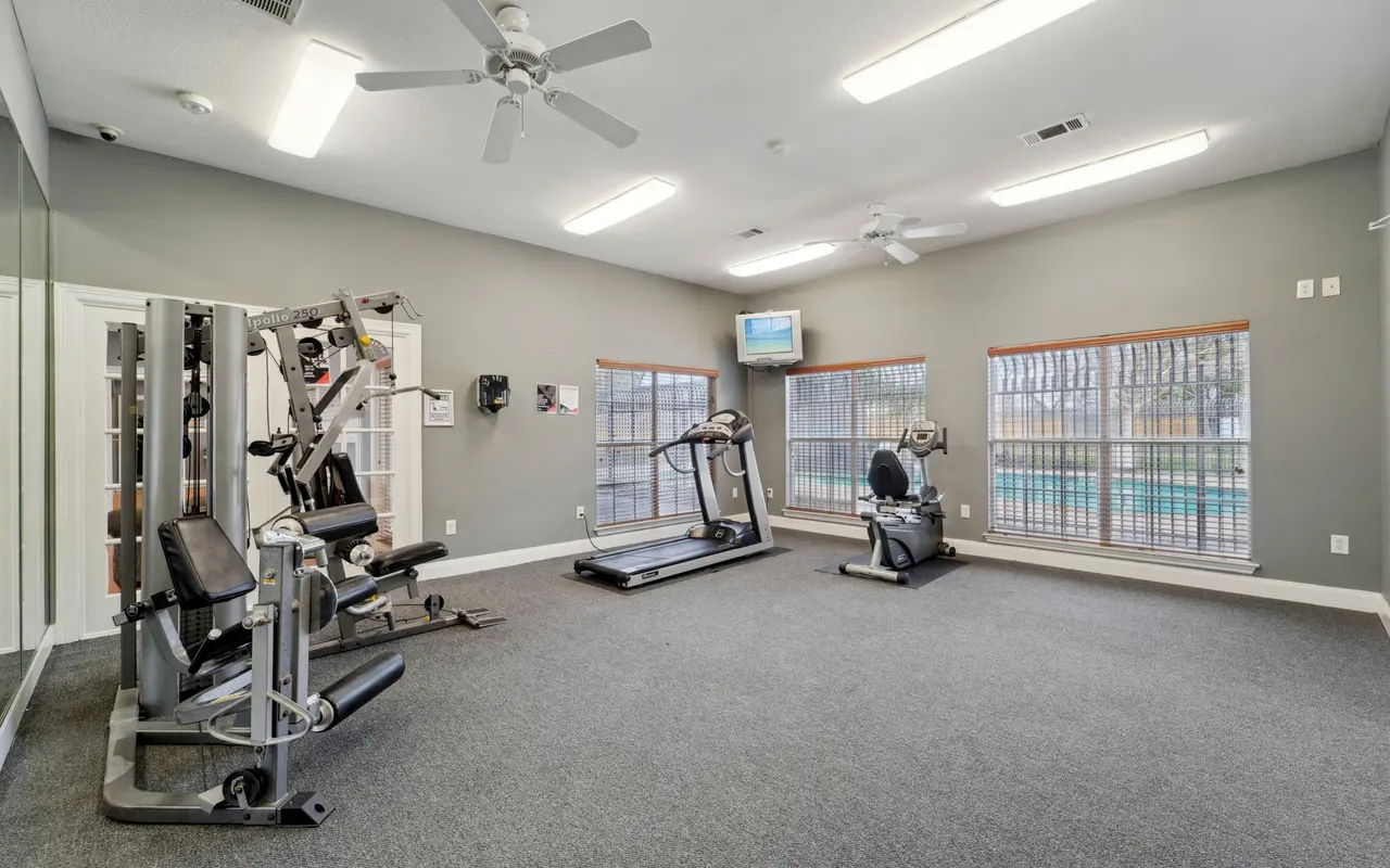 A fitness room featuring various exercise machines including a treadmill, an exercise bike, and a weight machine. Large windows provide a view outside, and there is a ceiling fan for ventilation.