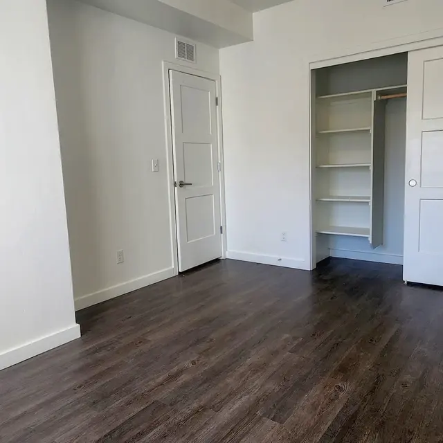 Canyon Terrace A spacious empty room with light-colored walls and dark wood flooring. There is a closet with shelves and a white door leading to another area. The lighting is bright, enhancing the openness of the space.