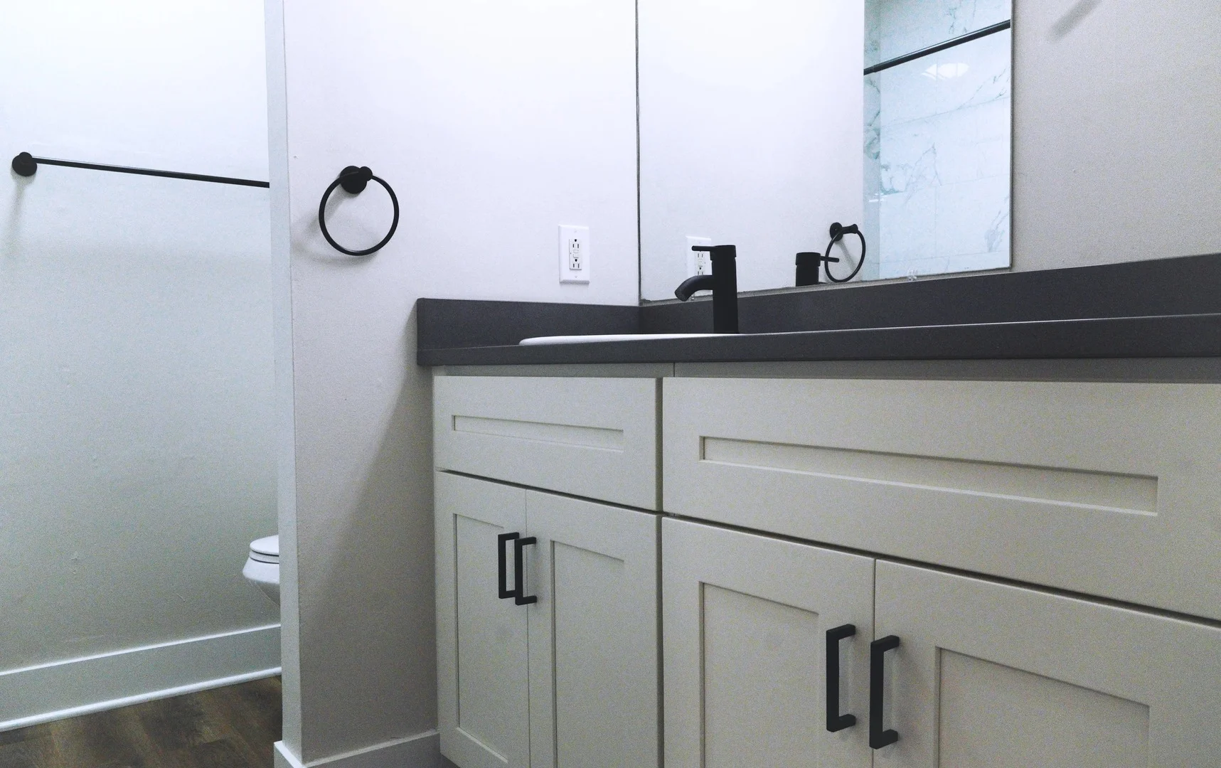 A modern bathroom featuring a sink with a dark countertop, cabinets with handle pulls, a wall-mounted towel rod, and a framed mirror. The bathroom has light-colored walls and wooden flooring.