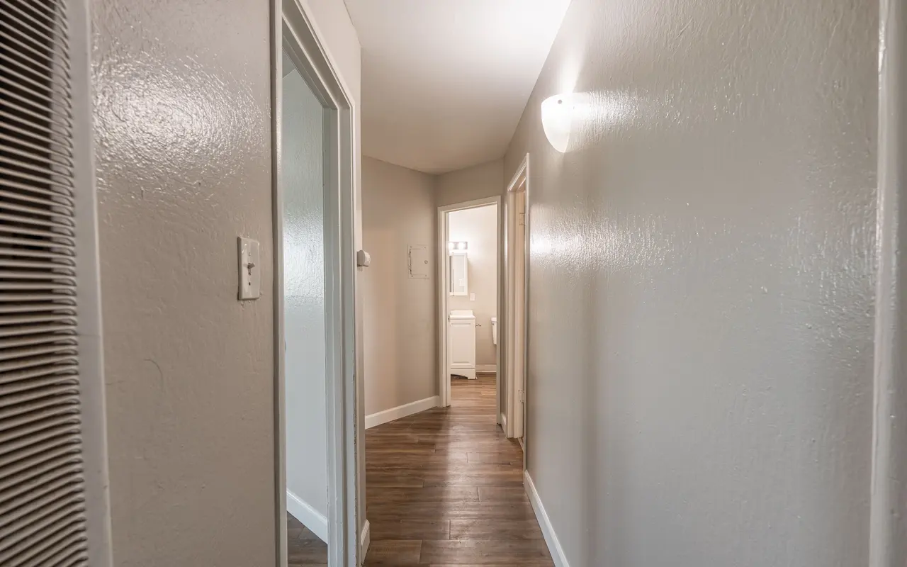 A long, narrow hallway with light-colored walls and a wood floor. Two doorways are visible on the left, and a bathroom can be seen at the end of the hallway. There is a wall-mounted light fixture illuminating the space.