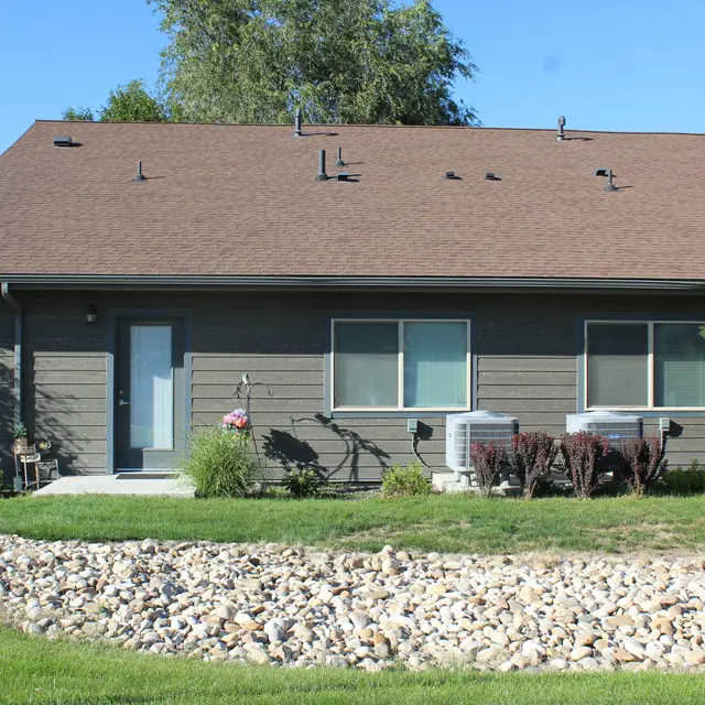A modern single-story house with a brown roof, featuring large windows and a front door. The landscaping includes a green lawn, decorative rocks, and small shrubs. Two air conditioning units are visible beside the house.
