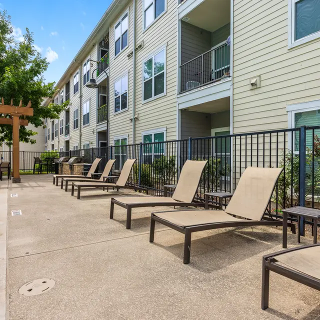 A well-maintained swimming pool area at an apartment complex, featuring sun loungers and a pergola. Green trees and shrubs are visible in the background next to the building.