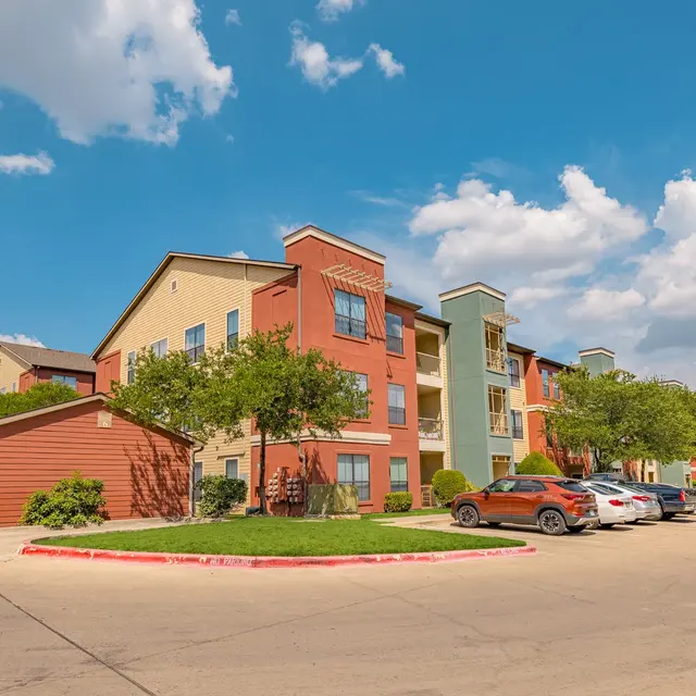 Modern Apartment Complex A modern apartment complex with colorful buildings surrounded by trees under a partly cloudy sky.  Parking area in the foreground with several cars.