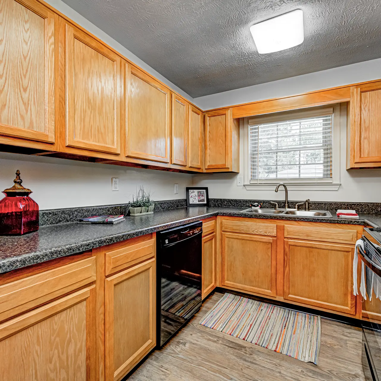 Modern Kitchen Interior A modern kitchen with wooden cabinets, a black countertop, and stainless steel appliances. The space features a sink under a window and a small decorative sign. A striped rug lies on the floor.
