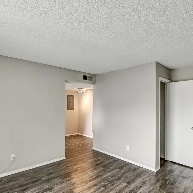 A vacant living room with light grey walls and laminate flooring. There is a closed door on the right and an open doorway in the background leading to another room.
