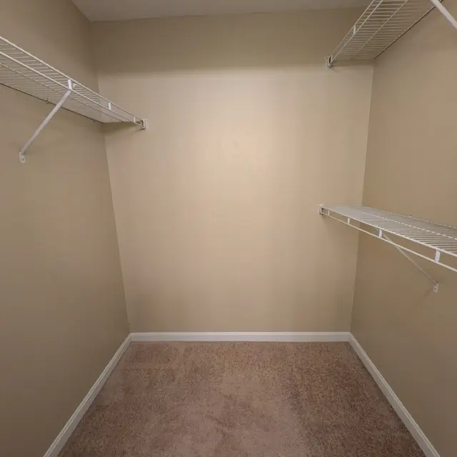 An empty closet with beige walls and carpet, featuring two wire shelves on opposite walls.