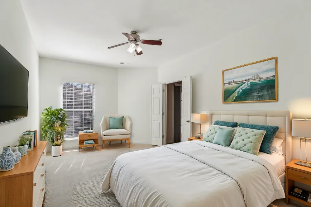A modern bedroom featuring a bed with white linens and teal accent pillows, a television mounted on the wall, and a plush chair next to a window. The room is illuminated with natural light and has decorative elements like a plant and artwork.