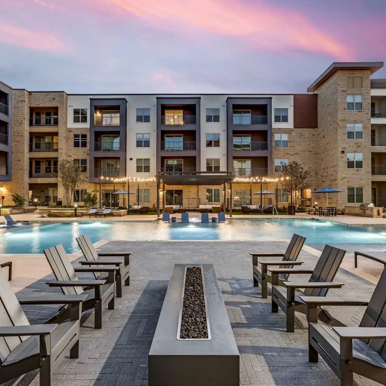 Modern Apartment Complex Pool Area at Sunset A modern apartment complex featuring a large swimming pool surrounded by lounge chairs and stylish outdoor furniture. The building has multiple floors and several balconies. The sky is painted with pastel colors during sunset.