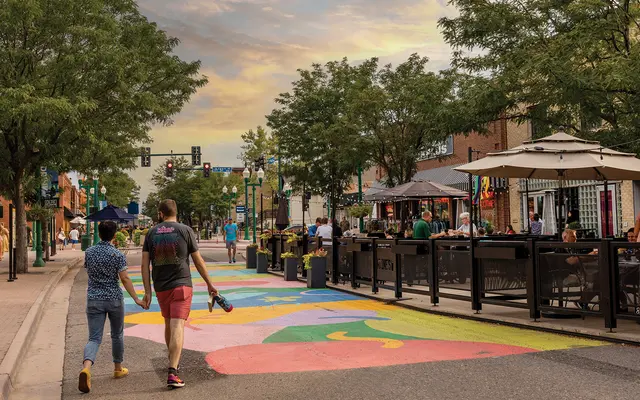 A couple walking hand in hand on a vibrant, colorful street surrounded by trees and outdoor seating areas. The sky is showing a sunset with soft clouds and a hint of color.