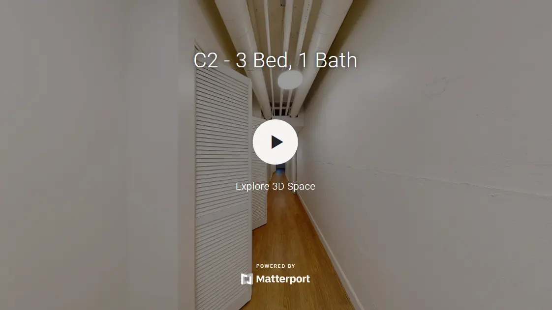 A narrow hallway leading to rooms, featuring white walls and a wooden floor. A video play button is centrally displayed, indicating a 3D space to explore.