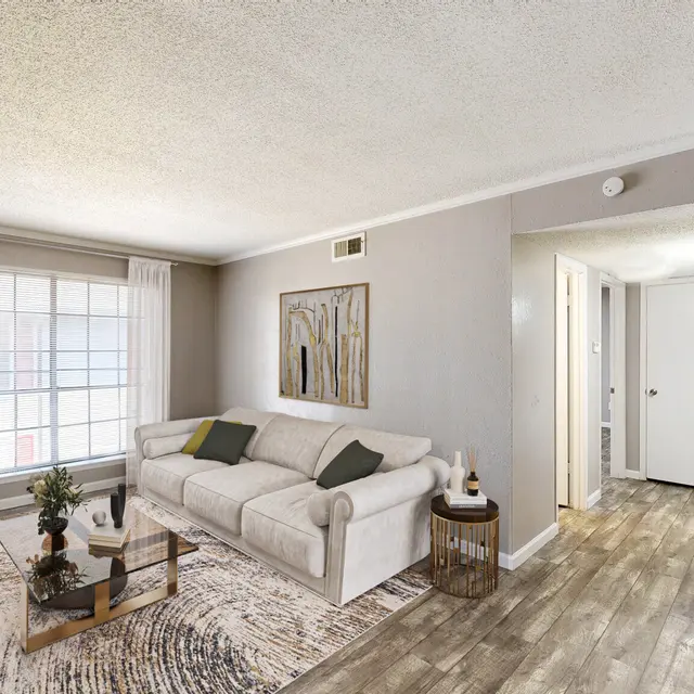 A modern living room featuring a light-colored sofa, two armchairs, and a coffee table arranged on a decorative rug. There are bookshelves on one side, a large window with white curtains, and an art piece on the wall. The flooring appears to be wood-like and the walls are painted in neutral tones.
