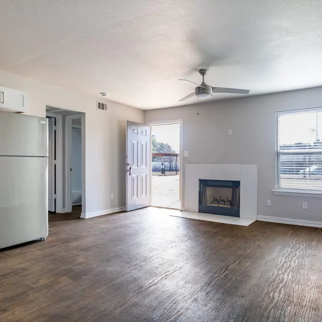 A spacious living area with wooden flooring, a kitchen area featuring a refrigerator and microwave, and a small fireplace. A door leads outside and a ceiling fan is visible.