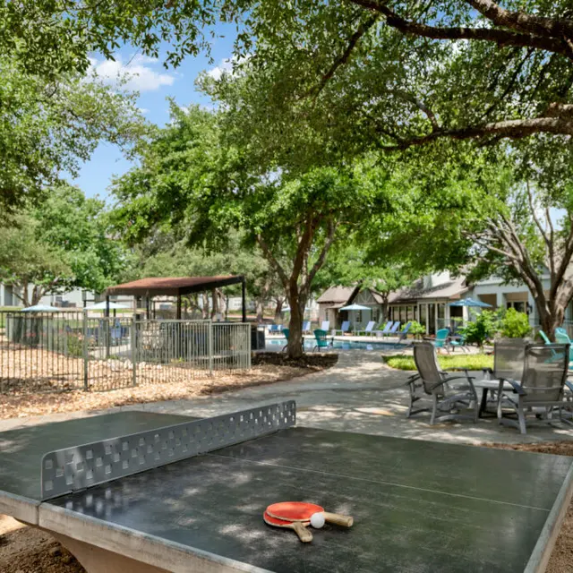 An outdoor area featuring a table tennis setup surrounded by trees and seating. A paddle and ball are placed on the table, with chairs and a swimming pool visible in the background.