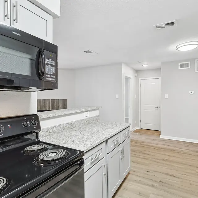 A modern kitchen featuring black appliances, granite countertops, and light wooden flooring. The kitchen includes a stove, microwave, and cabinetry with a clean layout and neutral-colored walls.