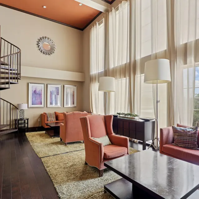A spacious modern living room featuring large windows with sheer curtains, a spiral staircase, and contemporary furnishings including orange armchairs and a dark coffee table on a light rug.