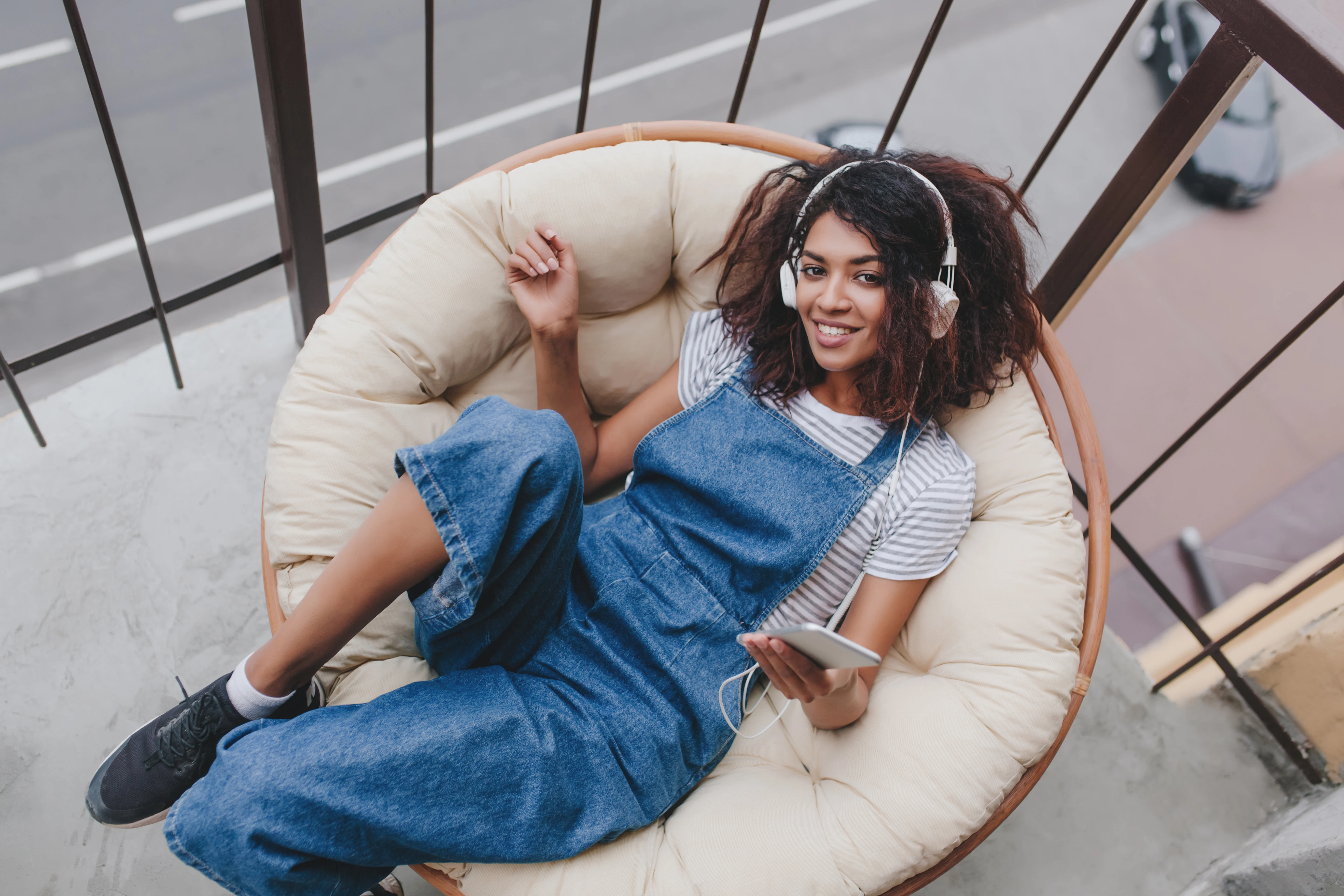 A young woman sitting comfortably in a round outdoor chair on a balcony, wearing denim overalls, a striped shirt, and headphones, smiling while holding a smartphone.