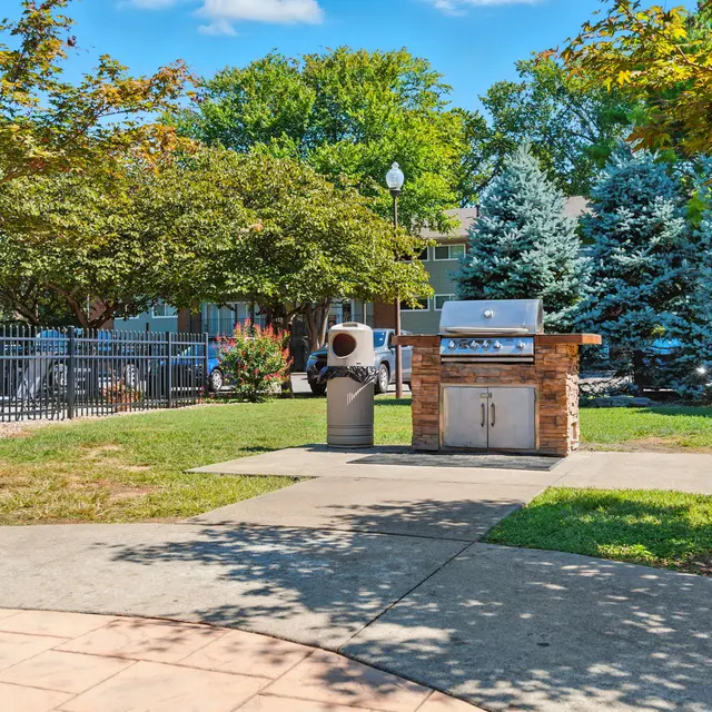 A sunny park grilling area featuring a stone barbecue grill, surrounded by green grass, trees, and two trash bins.