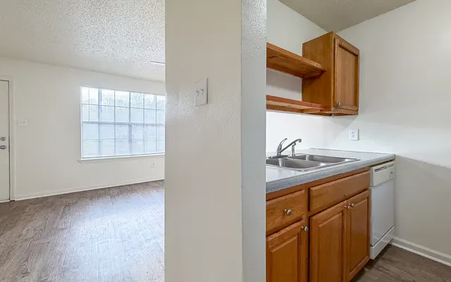A view of a small kitchen with wooden cabinets next to a living area with a large window.