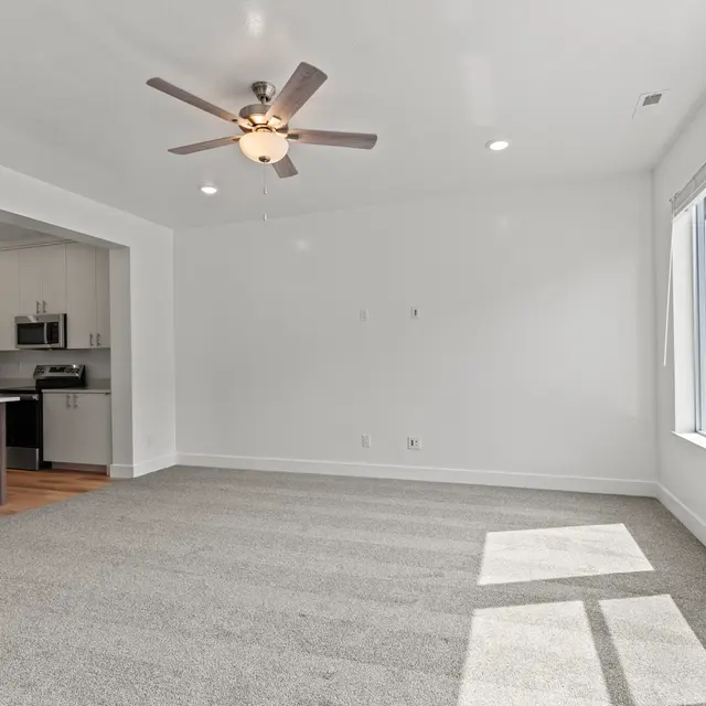 An empty living room with a ceiling fan and large windows letting in natural light. A modern kitchen is visible in the background with dark cabinetry and appliances.
