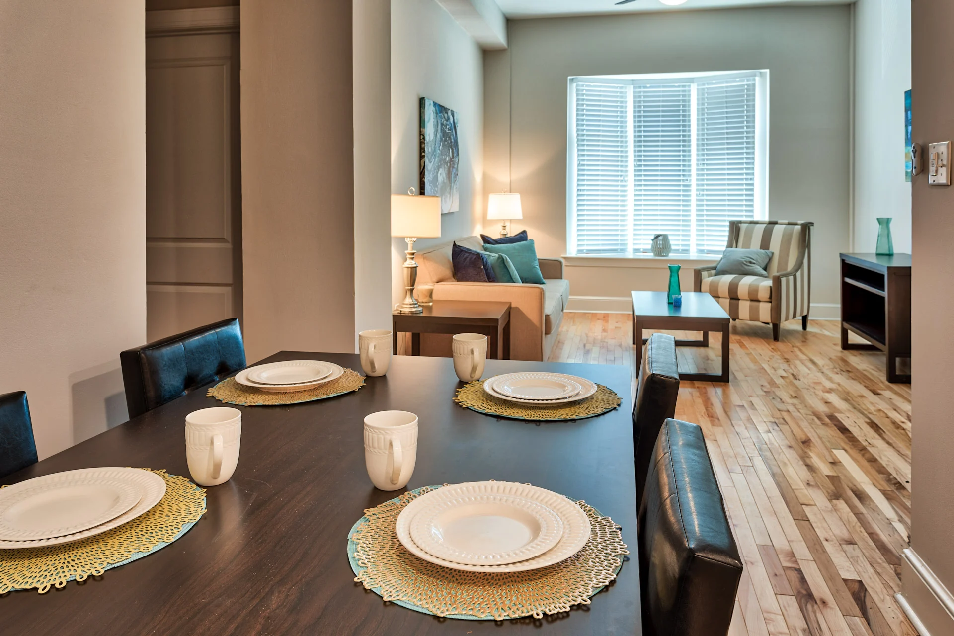Modern Dining and Living Area A modern dining area featuring a dark wooden table set for four with white plates and beige placemats. In the background, there is a bright living area with a couch, armchair, and large windows with white blinds.