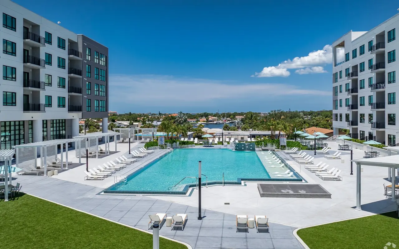 A spacious outdoor pool area surrounded by contemporary apartment buildings, featuring a large swimming pool, sun loungers, and green landscaping under a clear blue sky.