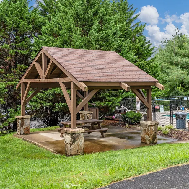 A wooden gazebo with a slanted roof, situated on a grassy area near a swimming pool, surrounded by green bushes.